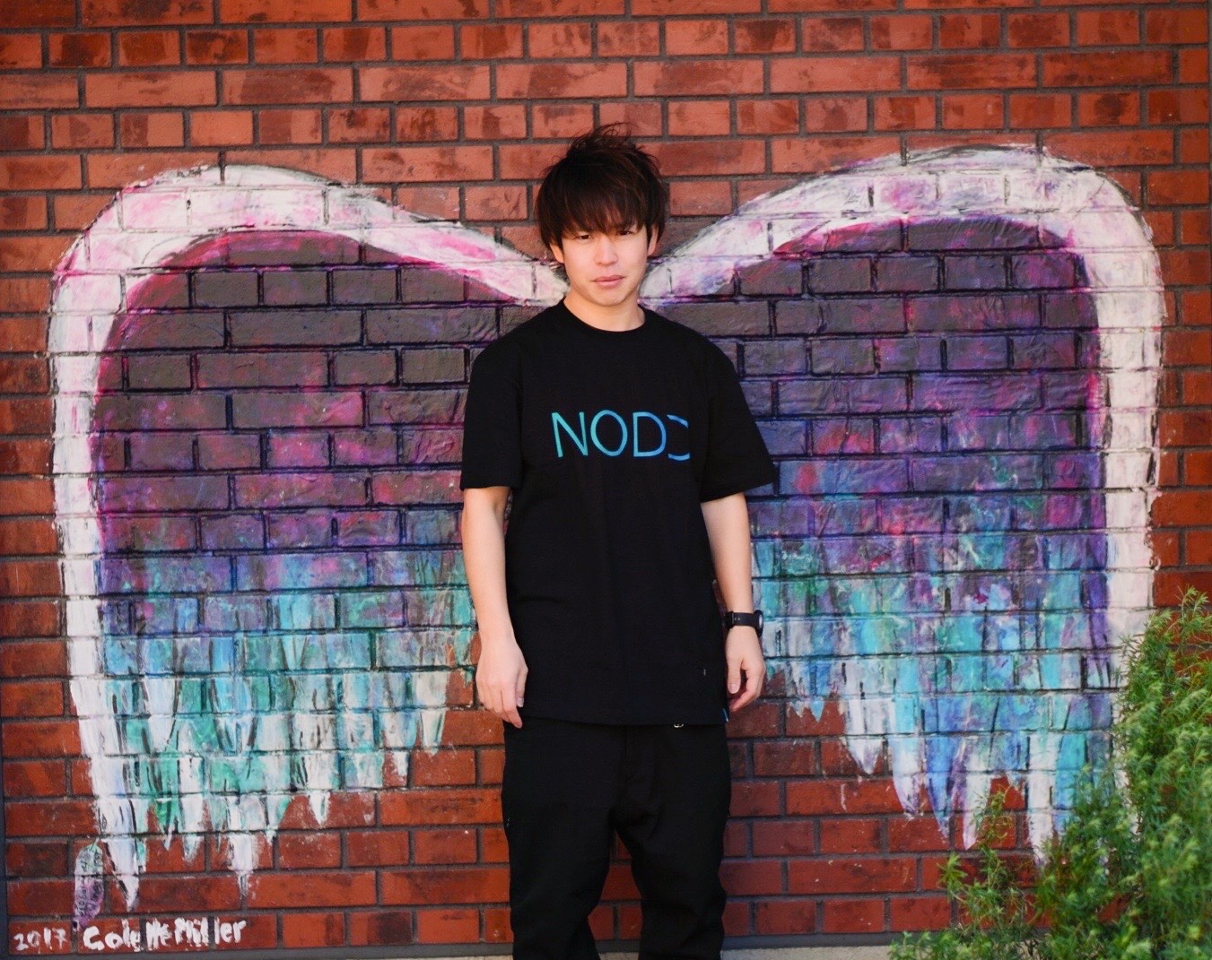NODDのTシャツで翼の壁画前に立つ石田（ステージ衣装にも合う引き立ち方）