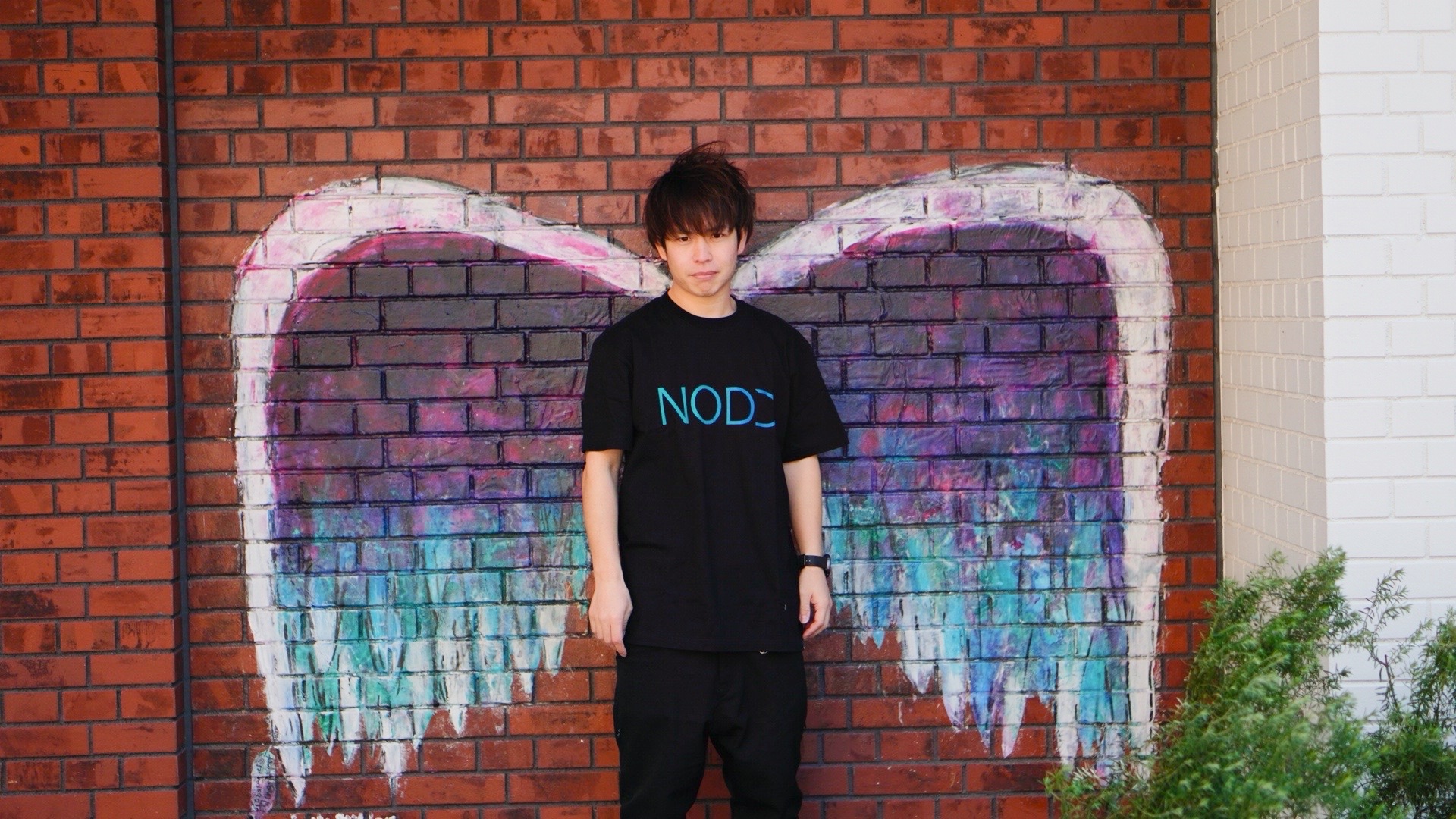NODDロゴTシャツ（黒）を着て翼の壁画前に立つ全身ショット